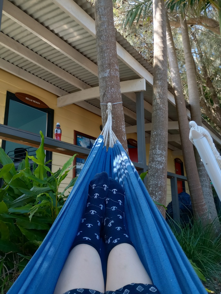Ein Bild von mir in einer blauen Hängematte. Das Bild ist aus meiner Perspektive aufgenommen, so dass man nur meine Beine und Füße sieht. Der Hintergrund ist mit einer der Surfcamp-Hütten und Bäumen geschmückt