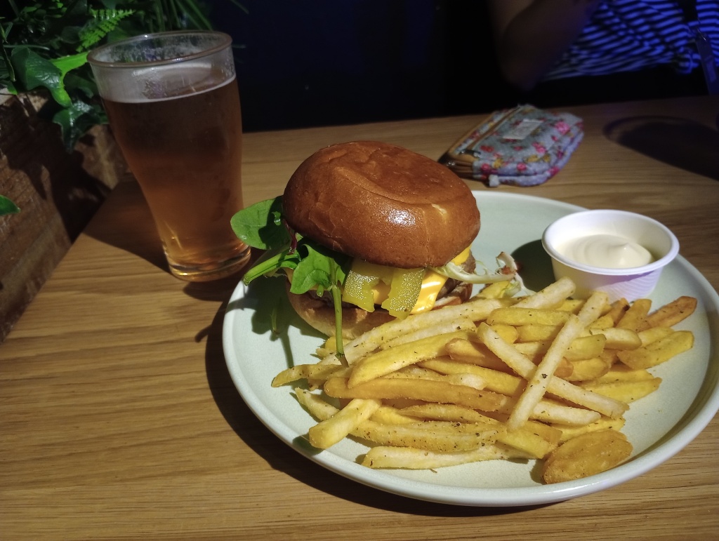 Ein BIld von meinem Essen auf einem Holztisch. Links steht ein Pint mit Bier, daneben ein großer Teller mit einem Burger, einer Portion Pommes und einem Schälchen Mayonnaise darauf.