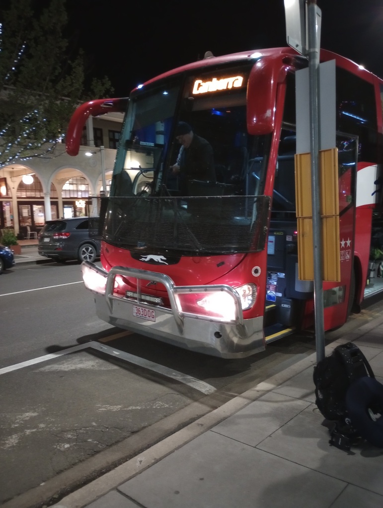 Ein Bild von einem roten Greyhound-Bus. Es ist nachts und er steht an einem gut beleuchteten Staßenrand in einer Stadt. Auf der Anzeigetafel oben am Bus kann man leicht verschwommen "Canberra" lesen