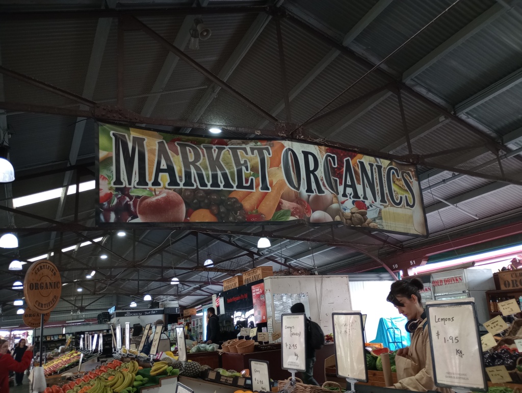 Ein Bild von einer Lagerhalle. Von der Decke hängt ein großes Banner mit der Aufschrift "Market Organics". Überall stehen stände mit frischem Obst und Gemüse
