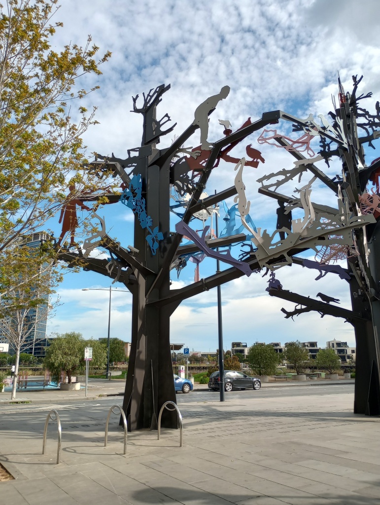 Eine Metall-Skulptur in Form eines Baums. Darafu sind weitere 2D-Metallmenschen angebracht, die auf dem Baum klettern. Im Hintergrund erstrahlt ein blauweißer Himmel